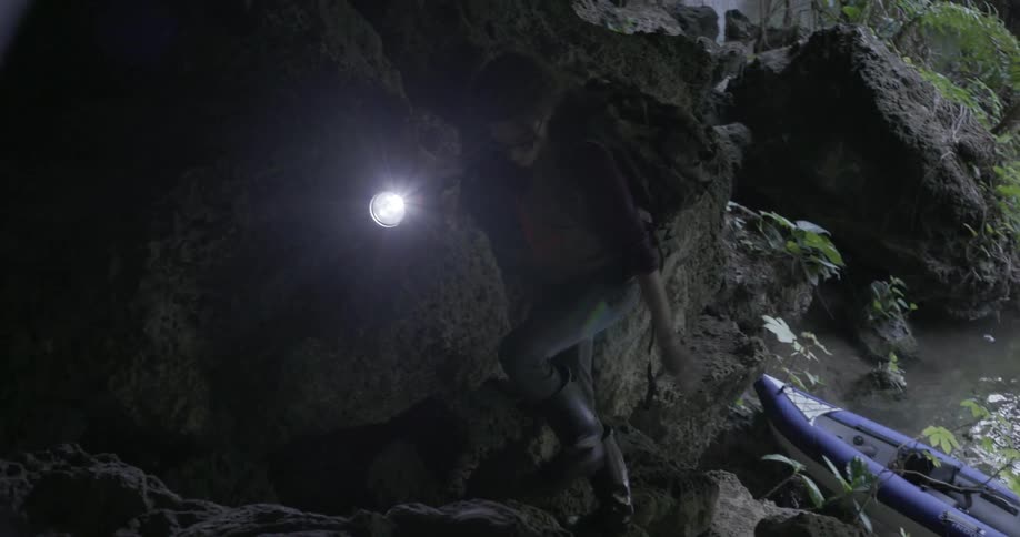 A woman carrying a flashlight climbs up a rocky cavern wall