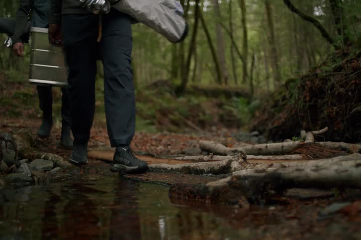 In slow motion, a group of hikers carefully crosses a makeshift log bridge over a stream in a dense, green forest