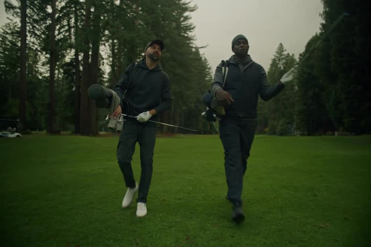 Two diverse male golfers walk in slow motion down the fairway of a lush, tree-lined course on an overcast day