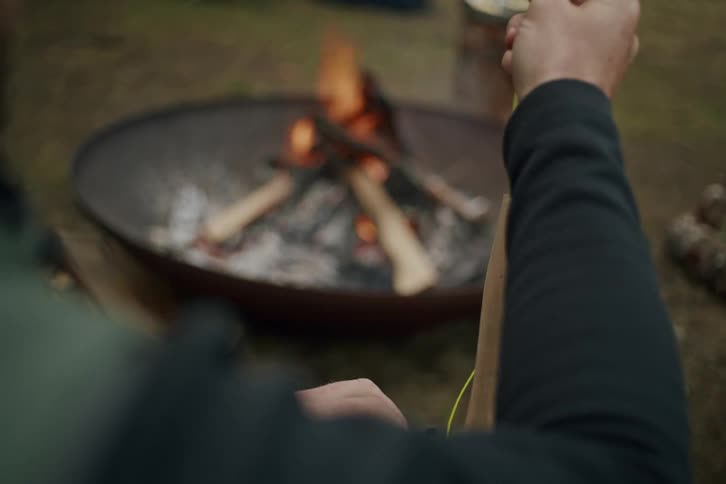 Over-the-shoulder view of a fisherman preparing his fly fishing rod next to a warm campfire