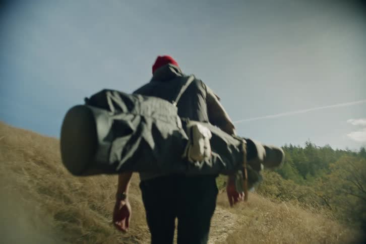 A slow-motion, low-angle shot follows a man in a red beanie carrying a large bag of gear as he hikes out of a forest and up a sunny, grassy hill