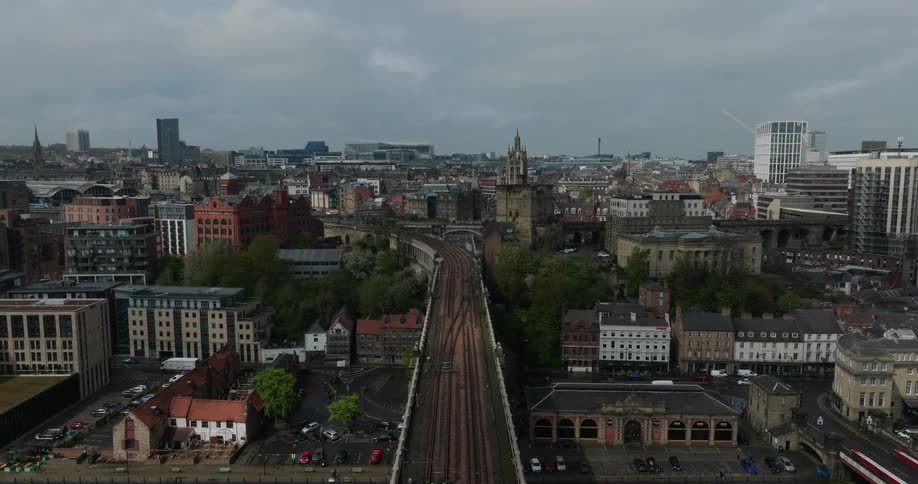 A slow aerial push-in shot follows a train traveling over the High Level Bridge into the city of Newcastle upon Tyne on a cloudy day