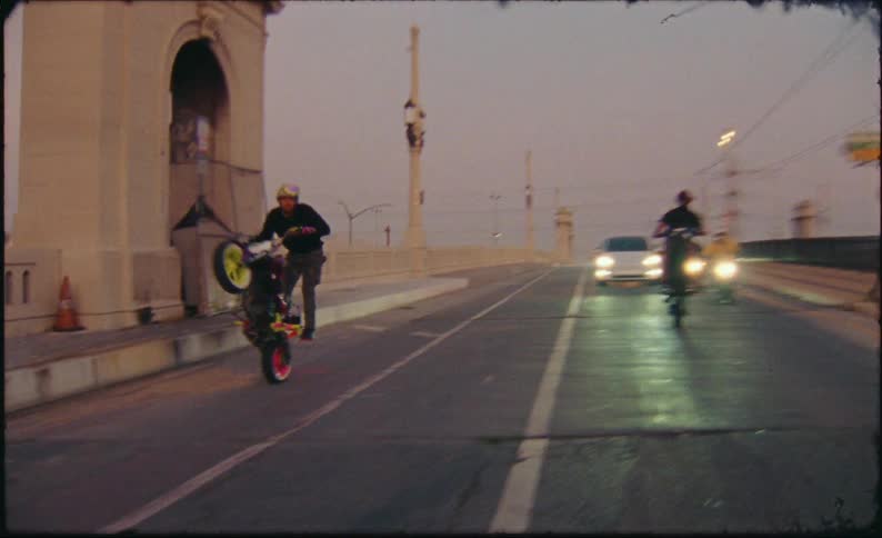 A biker gang expertly performs wheelies on their custom motorcycles as they ride across a city bridge at dusk in slow motion