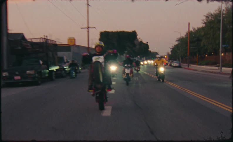 Vintage-style, slow-motion footage shows a man performing a long wheelie on his motorcycle, followed by his crew down a city street at dusk, creating sparks as he scrapes the ground