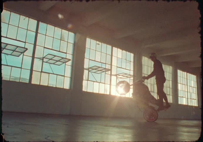 A vintage film shot captures a motorcycle stunt rider doing a wheelie in an empty warehouse at sunset