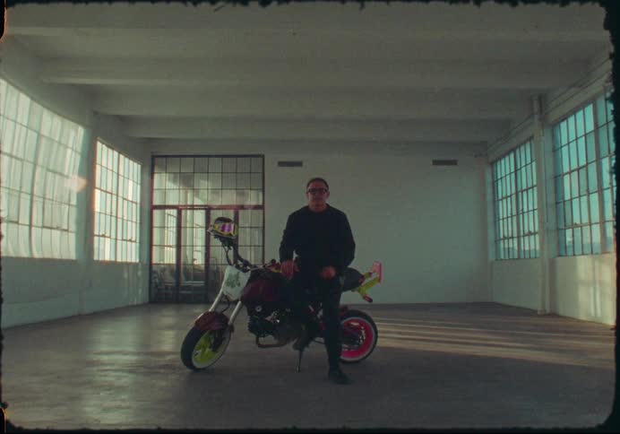 A man dressed in black poses with his colorful custom motorcycle inside an empty industrial warehouse with a vintage film look