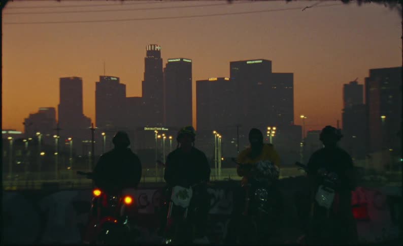 A gritty shot pulls back to reveal a group of silhouetted bikers on motorcycles against the Los Angeles skyline at sunset
