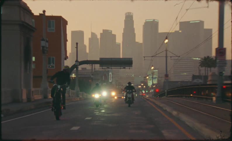 Motorcyclists perform wheelies on a bridge with the downtown Los Angeles skyline in the background during a hazy sunset