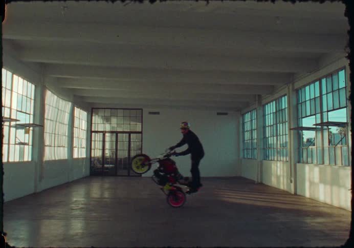 A skilled motorcyclist performs an impressive wheelie inside a vast, sunlit industrial warehouse on vintage-style film