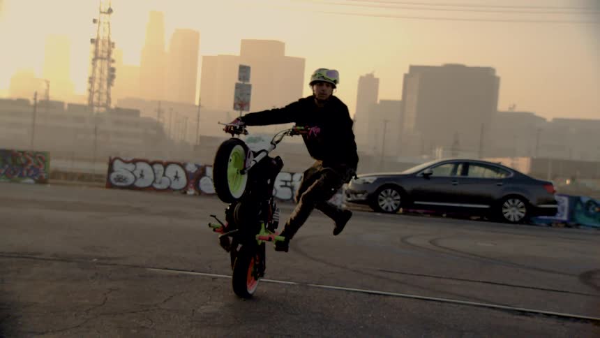 A skilled stunt rider performs a wheelie on a motorcycle in an urban lot, with the city skyline visible through the hazy sunrise