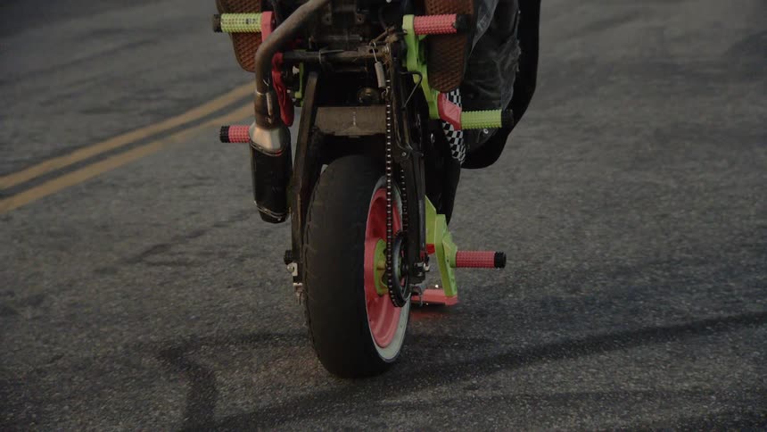 A stunt rider performs a wheelie on a custom mini bike, dragging a peg on the pavement and creating sparks in slow motion