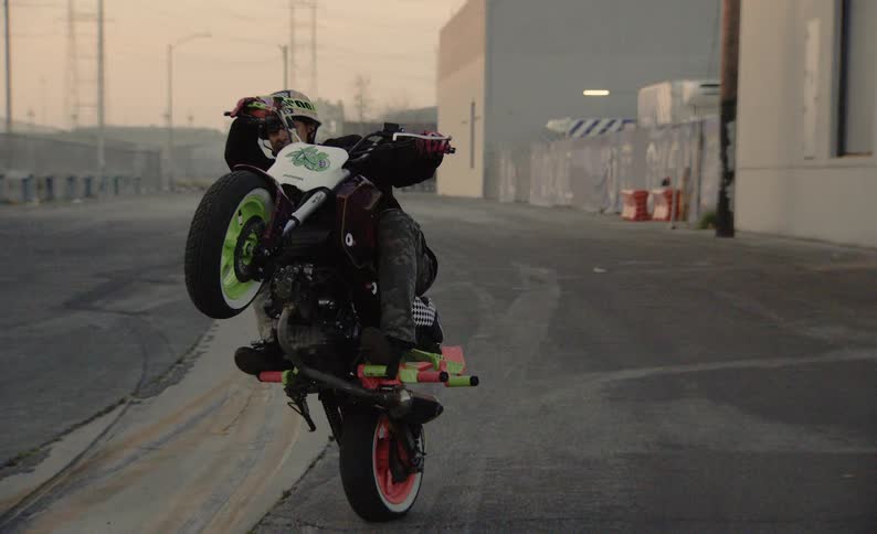 A stunt rider in slow motion holds a long wheelie on his motorcycle while riding down a gritty, industrial city street
