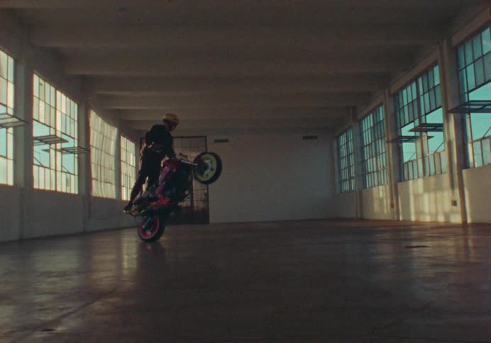 A silhouetted stunt rider performs a wheelie on a custom motorcycle inside a large empty warehouse as sunlight flares through the windows