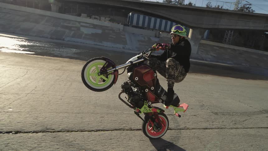 A stunt rider skillfully balances a colorful custom motorcycle in a wheelie within a sun-drenched urban drainage ditch