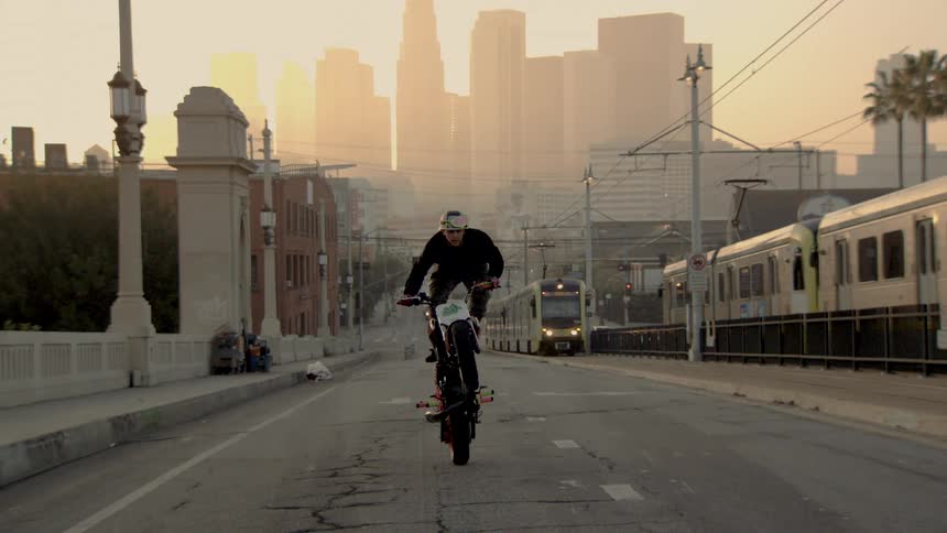 A stunt rider performs a wheelie on a motorcycle in slow motion on a city bridge with the Los Angeles skyline in the background at sunset