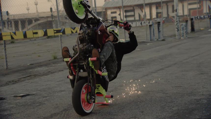 An extreme motorcycle stunt rider performs a static wheelie, dragging the tail and creating a shower of sparks in slow motion
