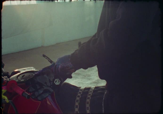 Vintage-style footage of a motorcyclist putting on a helmet and gloves before getting on a custom red bike in a garage