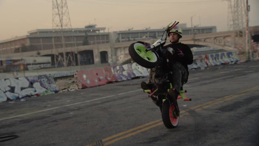 A skilled stunt rider performs a long, slow wheelie on a custom motorcycle in a gritty, urban landscape with graffiti-covered barriers
