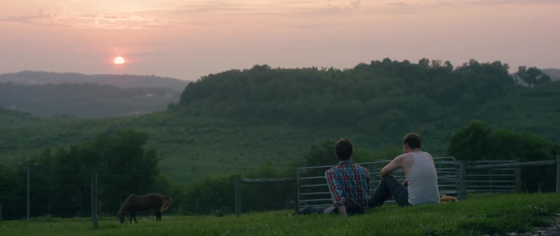 Two men and dog sitting on grass on ranch and talking at sunset