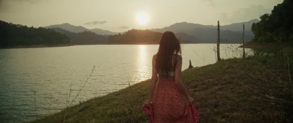 A woman in a flowing skirt walks in slow motion along a grassy bank, overlooking a serene lake and mountains at sunset