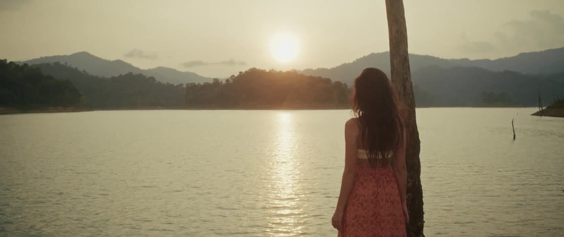 A woman in a flowing dress stands peacefully by a tranquil lake, watching the golden sunset over lush green mountains