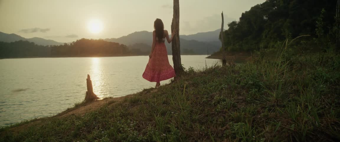 A woman in a long flowing skirt stands peacefully by a tranquil lake at sunset, admiring the scenic mountain view