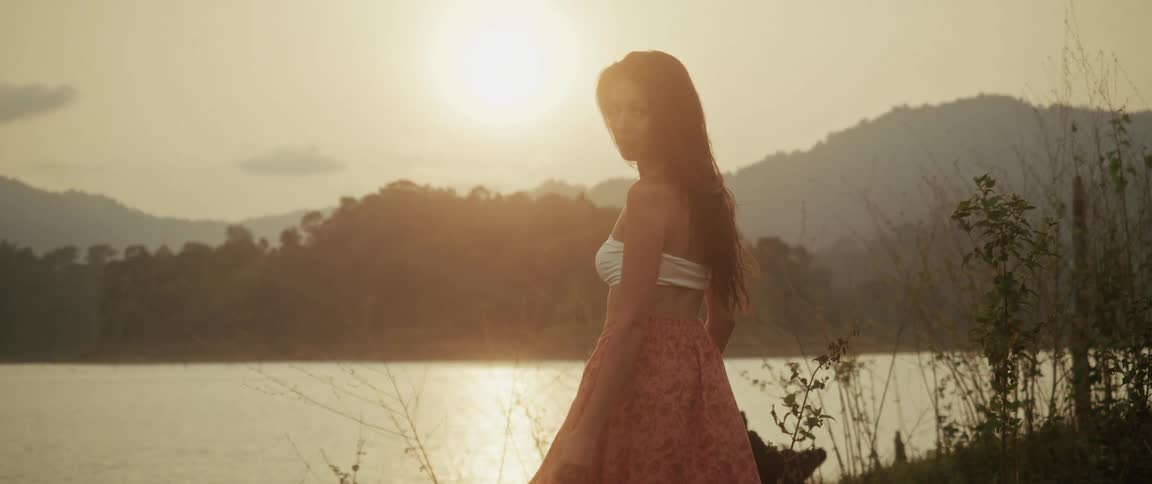 A woman in a flowing skirt enjoys the golden sunset by a tranquil lake, turning back to the camera in slow motion