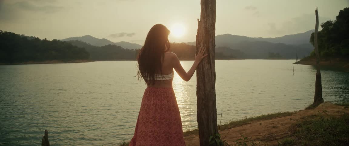 A beautiful woman in a flowing skirt leans against a tree and enjoys the golden sunset over a tranquil mountain lake