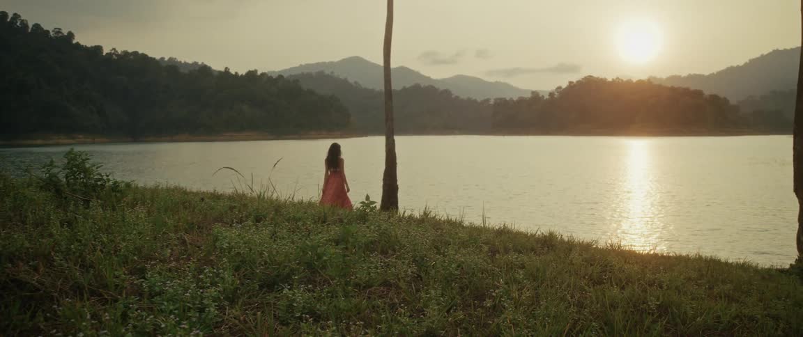 A woman in a long red dress walks to the shore of a tranquil mountain lake to watch the golden sunset