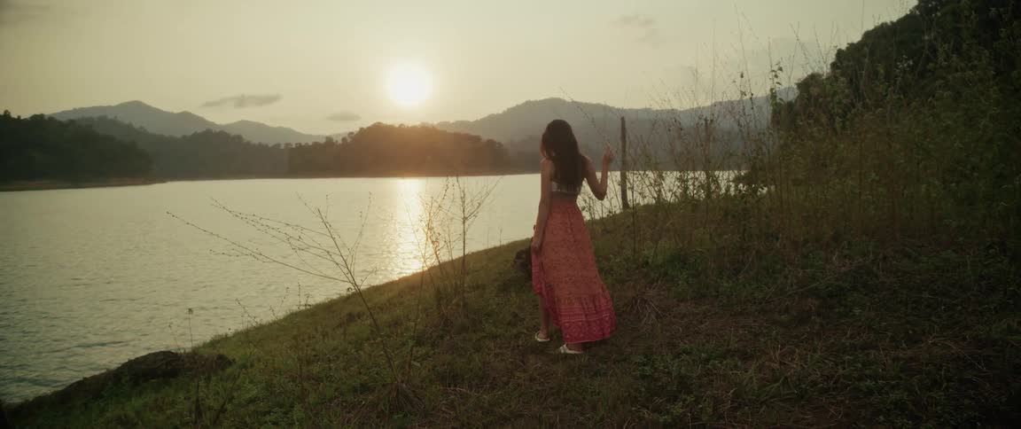 A beautiful woman in a flowing dress stands on a grassy bank overlooking a lake as the sun sets