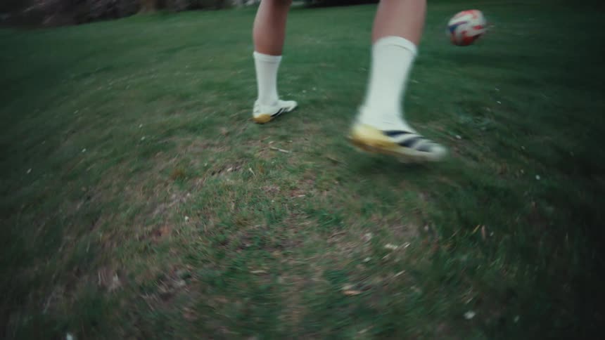 A dynamic, low-angle slow-motion shot follows a soccer player's feet as they skillfully dribble a ball across a grassy park