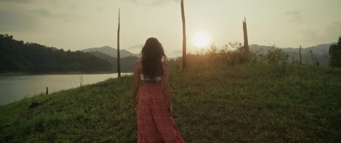 Slow motion shot of a woman in a long flowing skirt walking on a grassy hill by a lake, turning to smile at the camera during a golden sunset