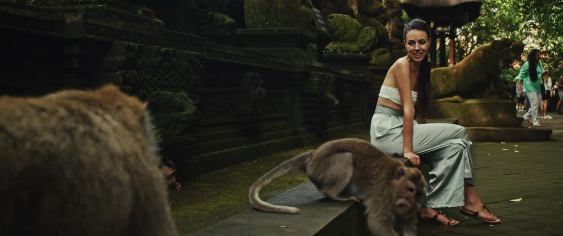 A smiling woman watches a family of wild macaque monkeys at a mossy jungle temple in slow motion