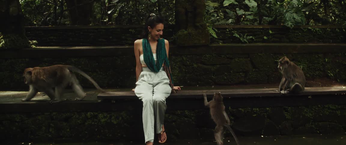 A happy woman sits on a bench in a lush green jungle park, smiling as she is surrounded by several playful monkeys