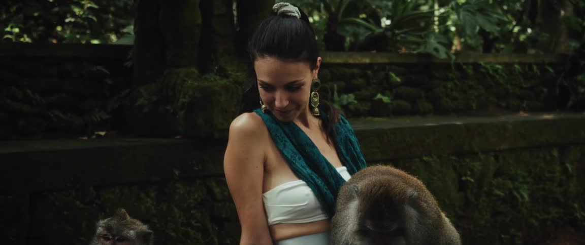A woman smiles while a monkey rests peacefully on her lap as she sits on a bench in a lush, green jungle