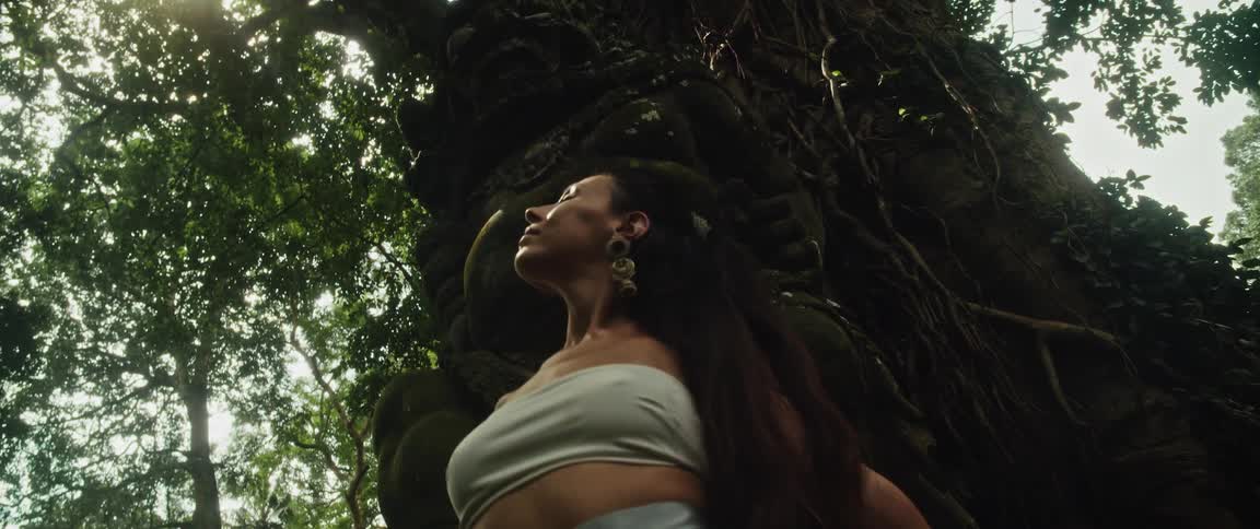 A woman in a serene, meditative pose leans against an ancient moss-covered deity statue in a lush jungle as the camera pans up to reveal the sunlit canopy