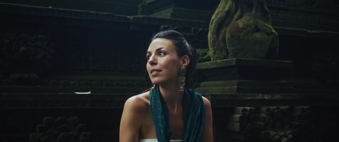 A serene woman with a turquoise scarf smiles peacefully while sitting amidst the ancient, moss-covered stone ruins of a temple