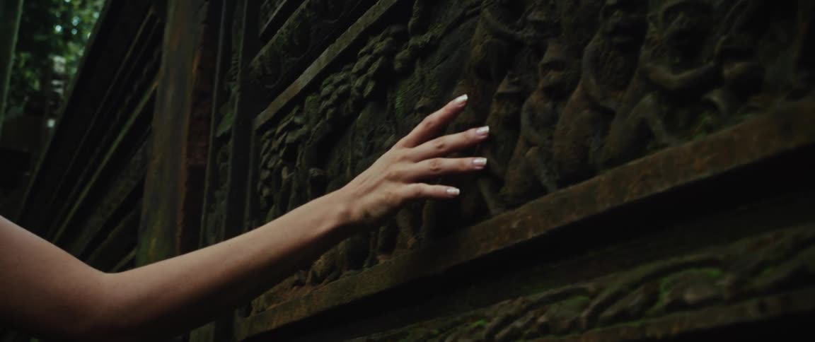 A woman's hand gently slides across an ancient, moss-covered stone bas-relief at a temple