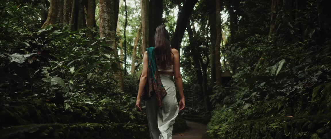 A graceful woman dressed in flowing clothes walks in slow motion down a stone path into a lush and mystical green jungle