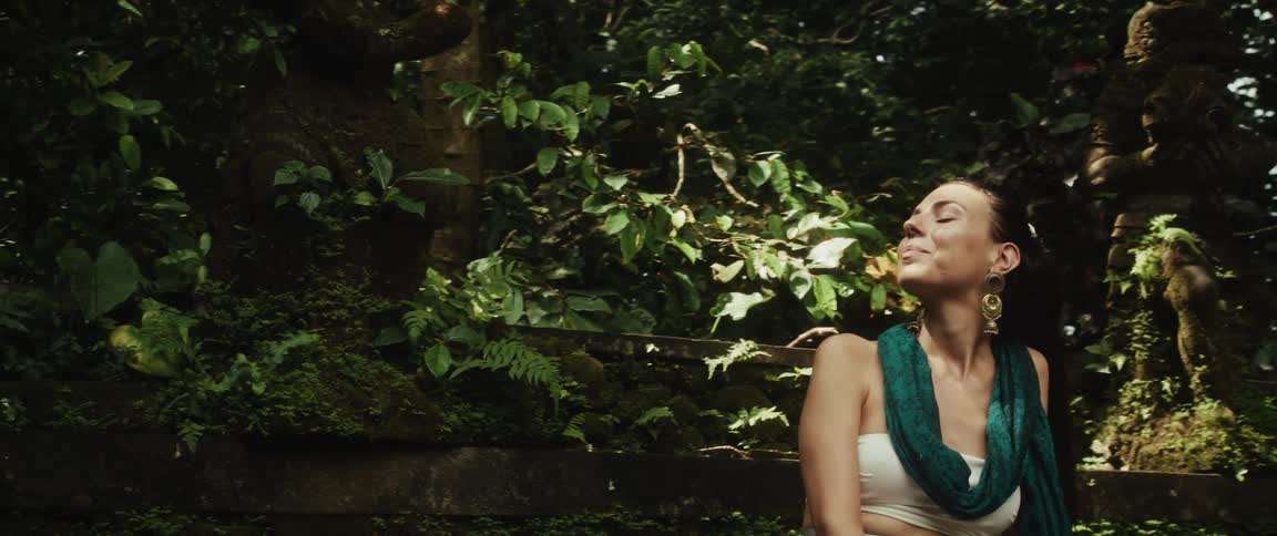 A smiling woman sits peacefully on a bench next to a grooming monkey in a lush, green jungle sanctuary