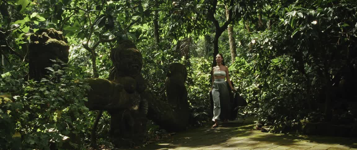 A woman in elegant clothing walks through a tranquil jungle path past ancient moss-covered statues