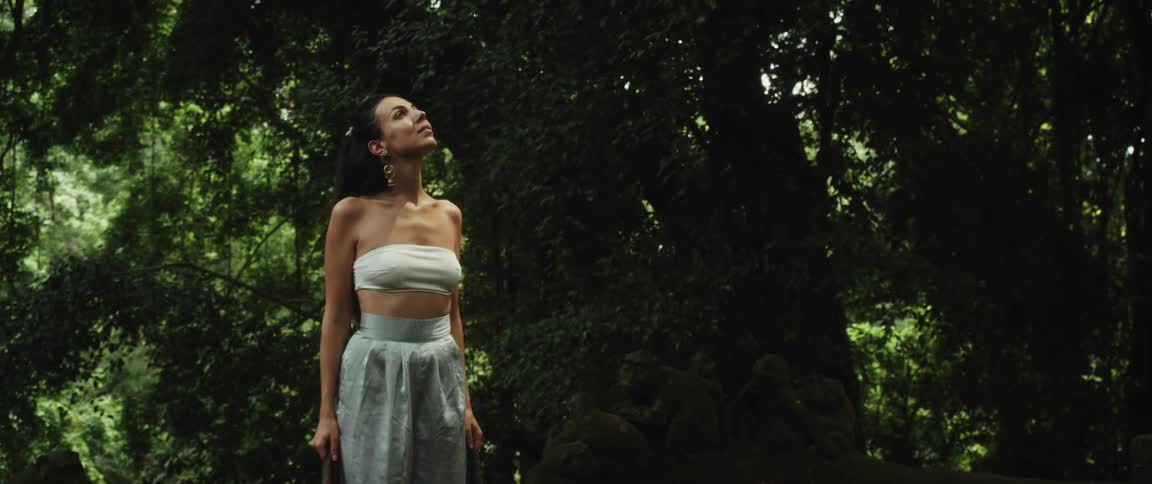 A woman peacefully explores a lush, green jungle, walking amongst ancient, moss-covered stone statues