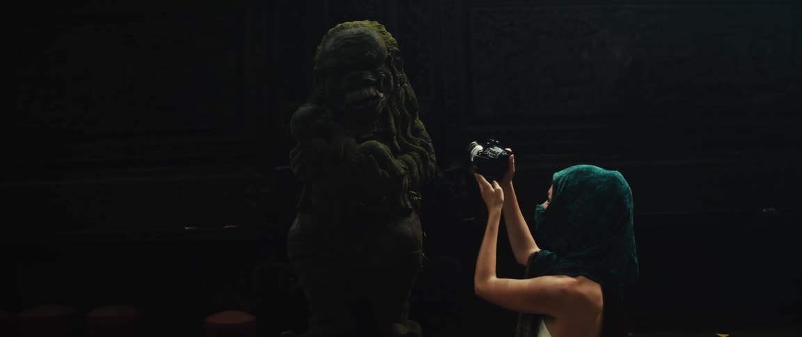 A woman in a green head covering takes a slow-motion photo of an ancient, moss-covered statue in a dark temple