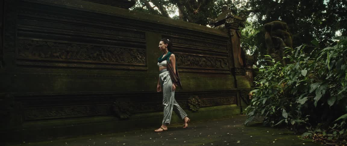 A woman in flowing clothes peacefully explores an ancient, moss-covered stone temple with intricate carvings deep within a lush jungle