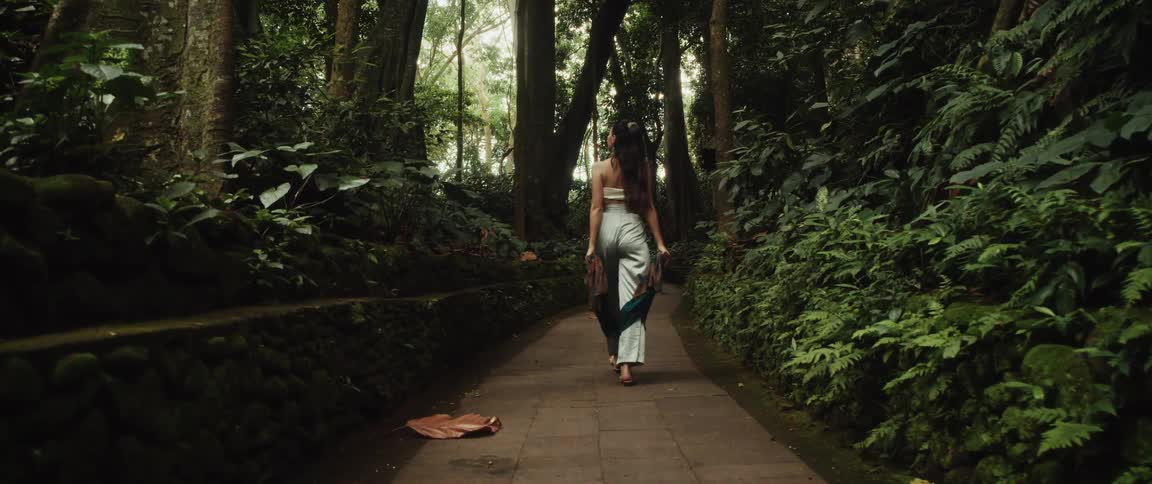 A woman gracefully walks barefoot down a stone path in a lush tropical jungle, her colorful scarf trailing behind her in slow motion