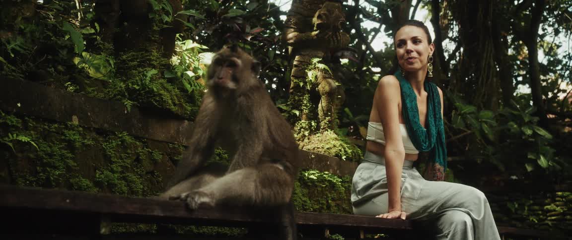 A smiling woman sits peacefully next to a large macaque monkey on a bench in a lush, tropical jungle sanctuary