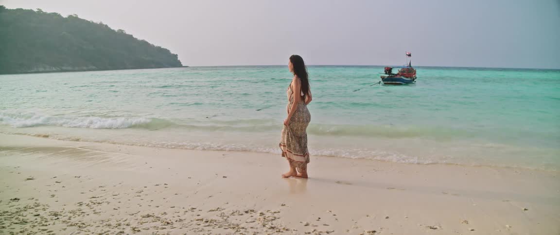 Slow motion shot of a woman in a flowing dress walking barefoot along a tranquil tropical beach next to the calm, turquoise ocean
