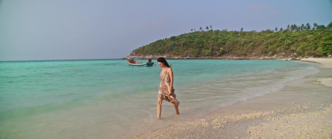 A woman in a flowing dress walks barefoot along the shoreline of a pristine tropical beach in slow motion
