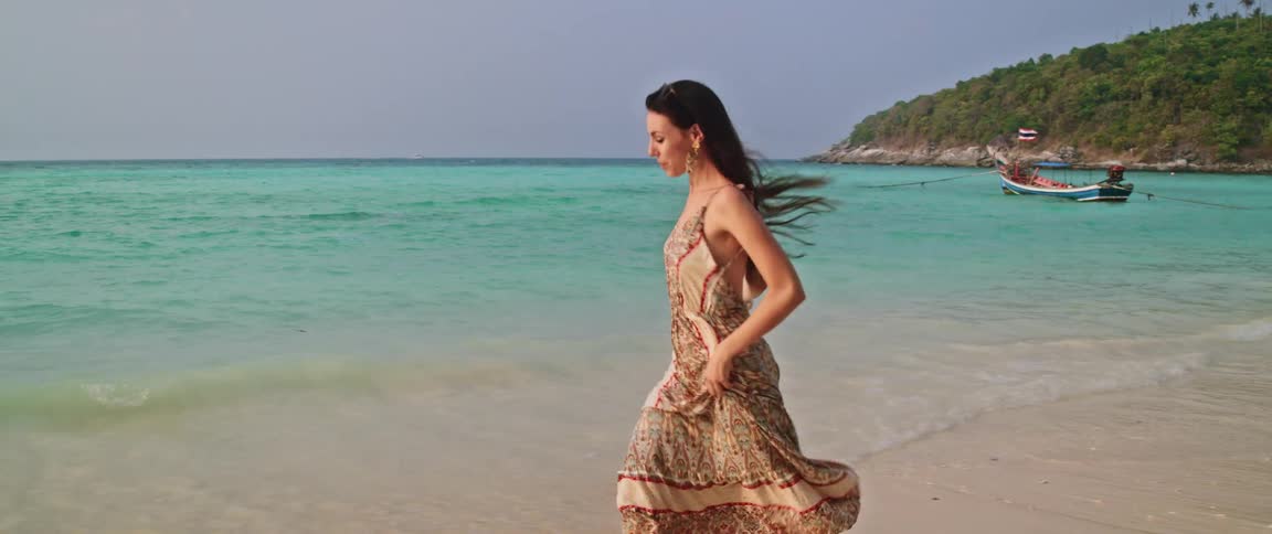 Slow motion shot of a smiling woman in a long dress running freely on a beautiful sandy beach next to the turquoise ocean