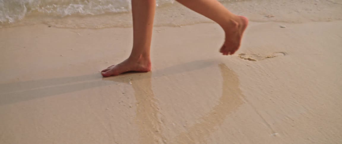 A slow-motion close-up shot of a person's bare feet walking along a sandy shoreline as gentle waves wash over them